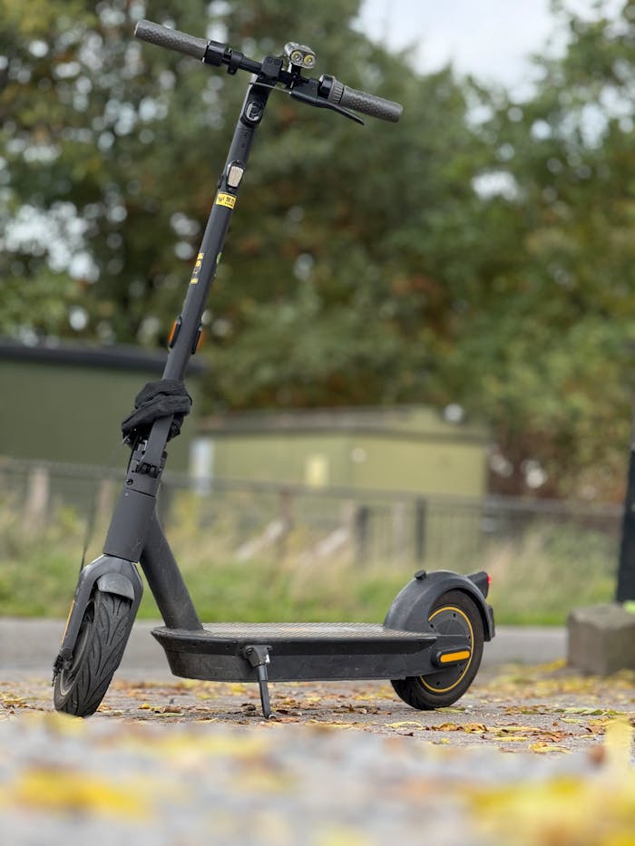 A sleek black electric scooter parked outdoors on a path covered with autumn leaves.